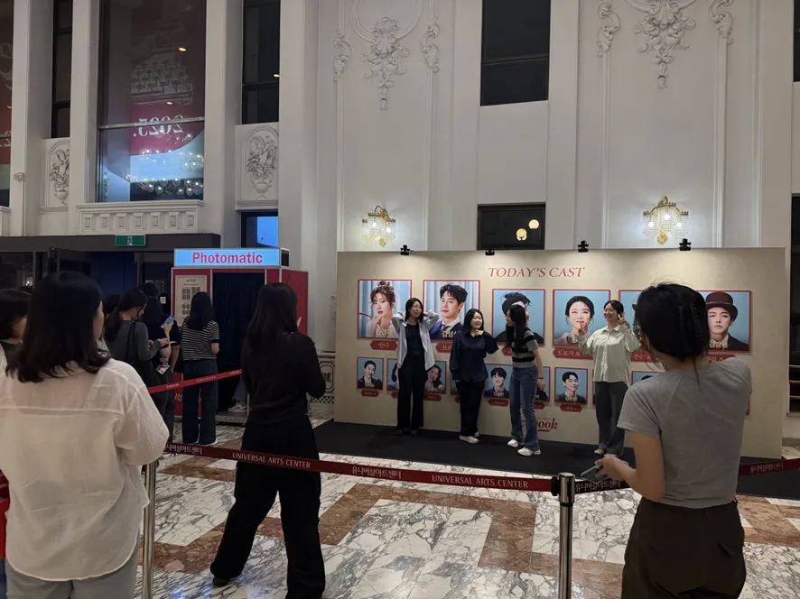 thumbnail - 서울 광진구 유니버설아트센터 로비에 설치된 ‘TODAY’S CAST’ 포토존. 관객들이 기념 촬영을 하고 있으며, 인생네컷 형태의 포토부스도 운영된다.