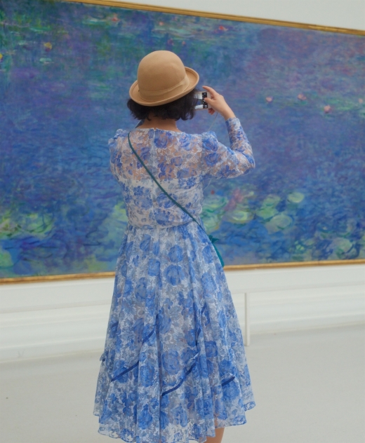 파스텔 색감이 특징인 클로드 모네의 한 작품 앞에서 카메라를 들고 사진을 찍고 있는 여성은 작품에 동화된 것처럼 푸른색 파스텔 색상의 꽃무늬 원피스를 입고 있다.