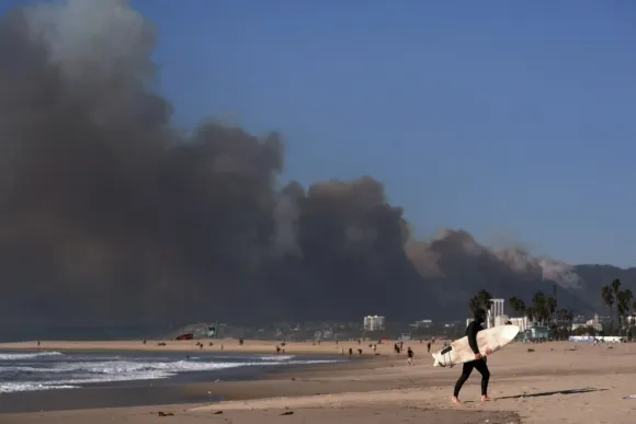 7일 오전(현지시간) 미국 캘리포니아주 로스앤젤레스의 퍼시픽 팰리세이즈에서 대형 산불이 발생했다. 짙은 연기로 뒤덮인 해변을 걷는 서퍼의 모습.  AP 연합뉴스